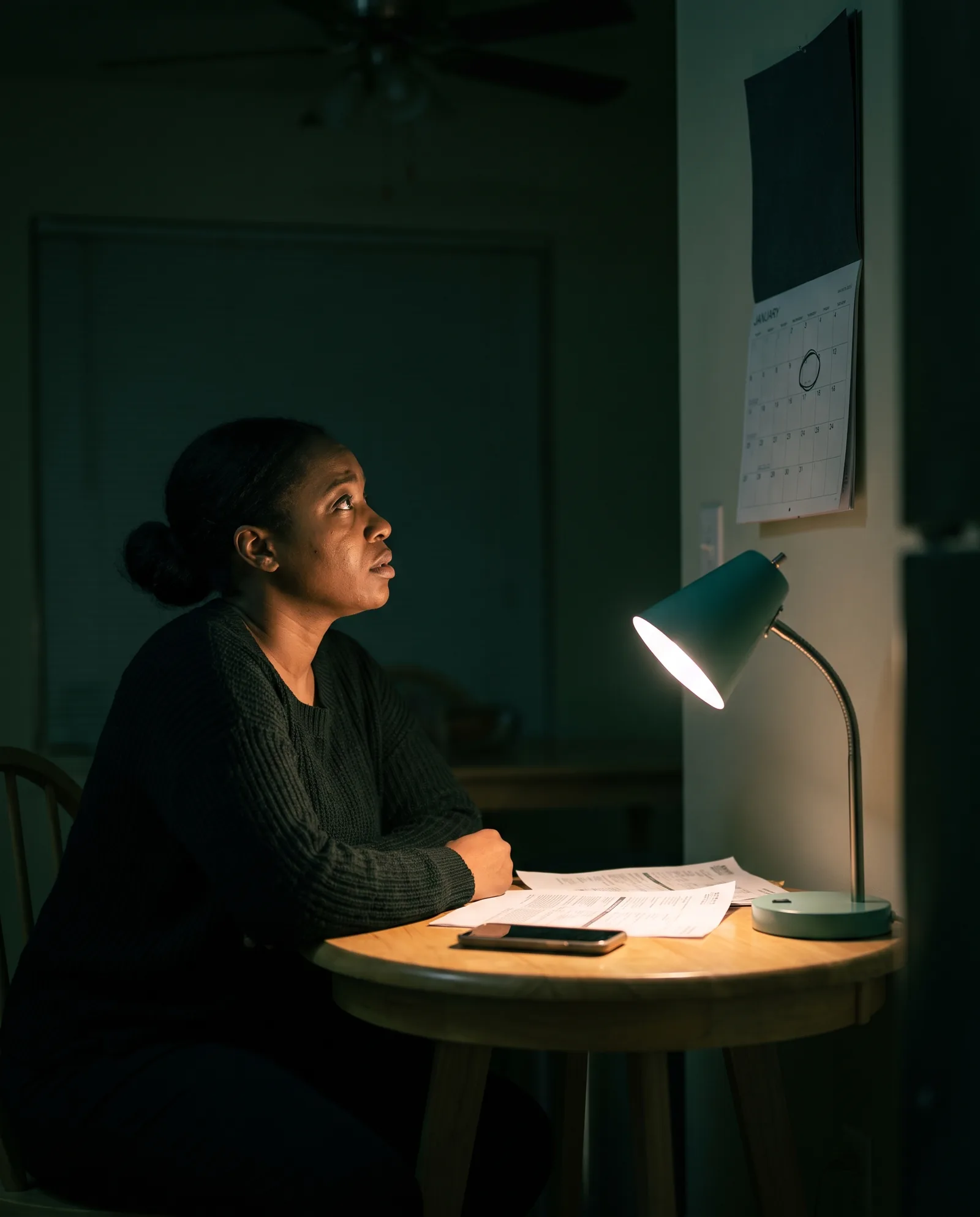 A woman at a small table at night under a lamp, court papers in front of her, looking up at a calendar