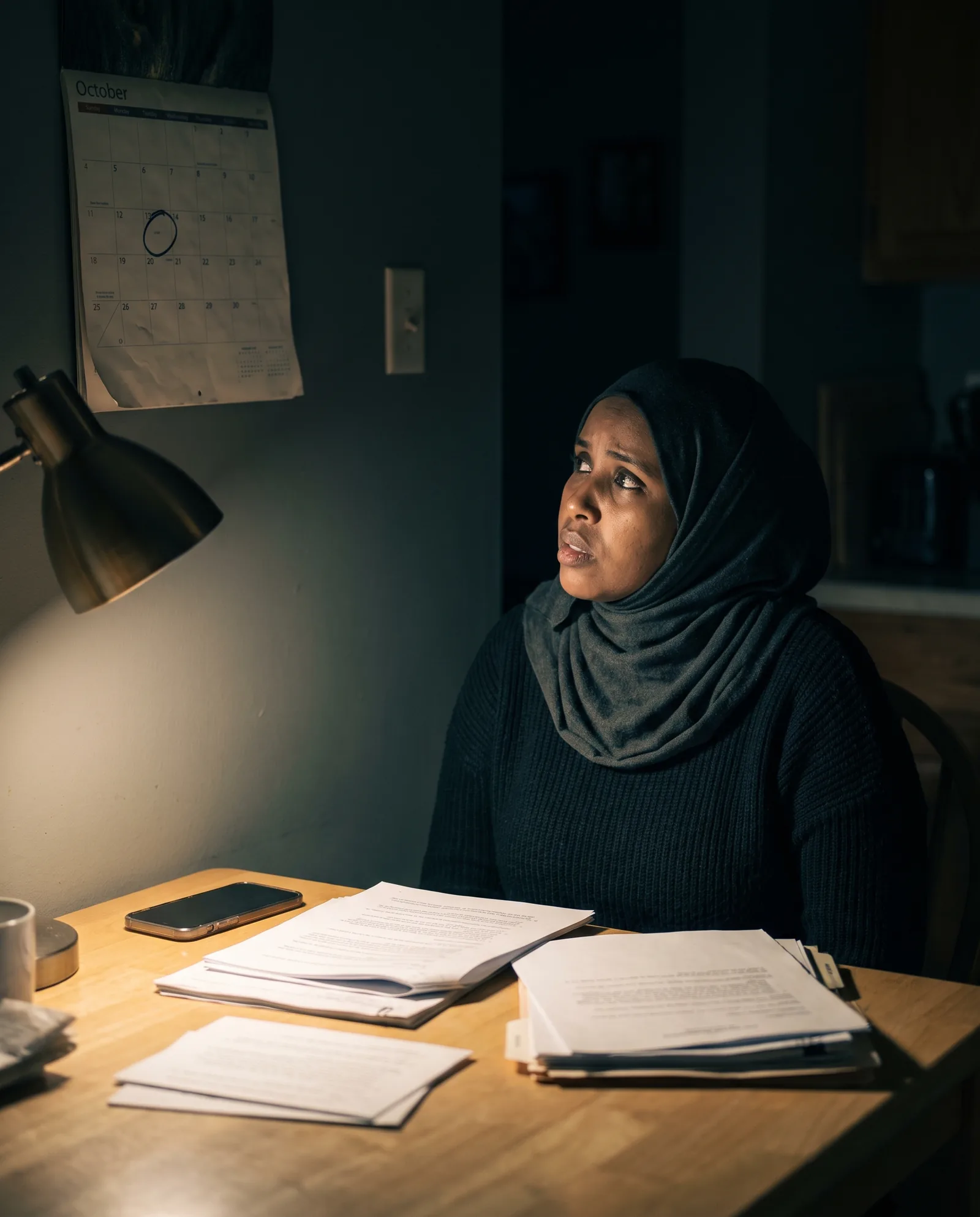 A woman in a headscarf at a small table at night under a lamp, court papers in front of her, looking up at a calendar