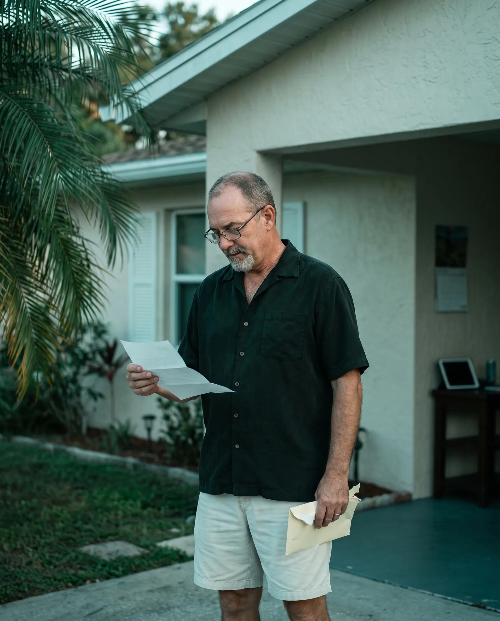 A man outside a pale stucco Florida house with palms, reading a letter he has just opened, calm